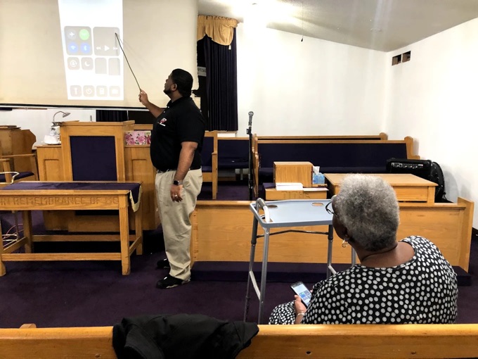 A TechEd and Solutions instructor teaching a phone class in a church, pointing at a projected phone screen while an attendee follows along on her own phone